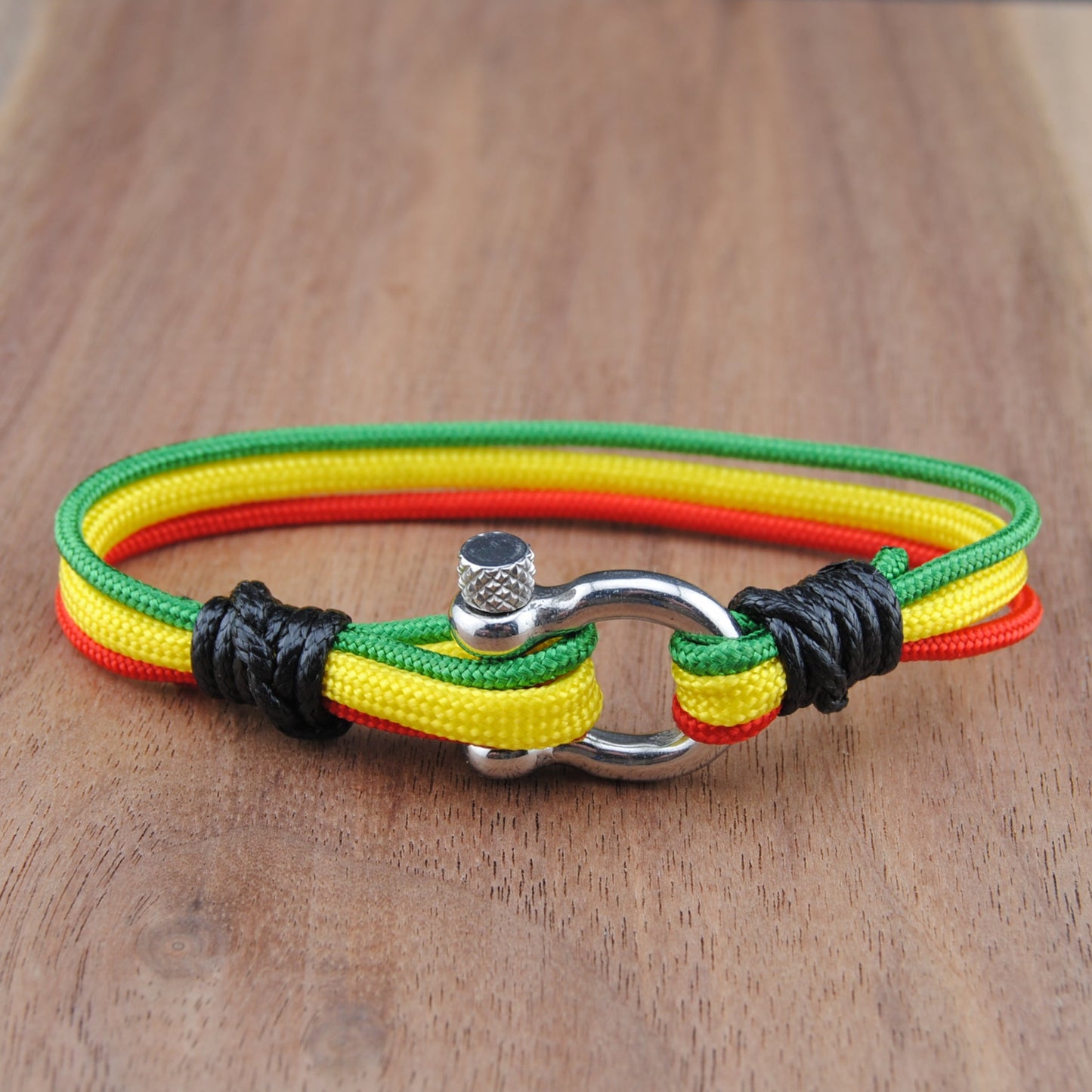 A multi-strand cord bracelet with green, yellow, and red stripes, featuring black knotted accents and a silver metal clasp, displayed on a wooden surface.