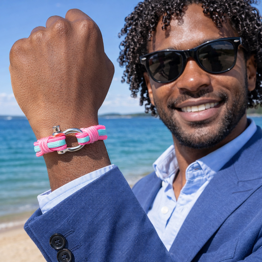 Man on a beach wearing sunglasses and a blue jacket with a pink bracelet.