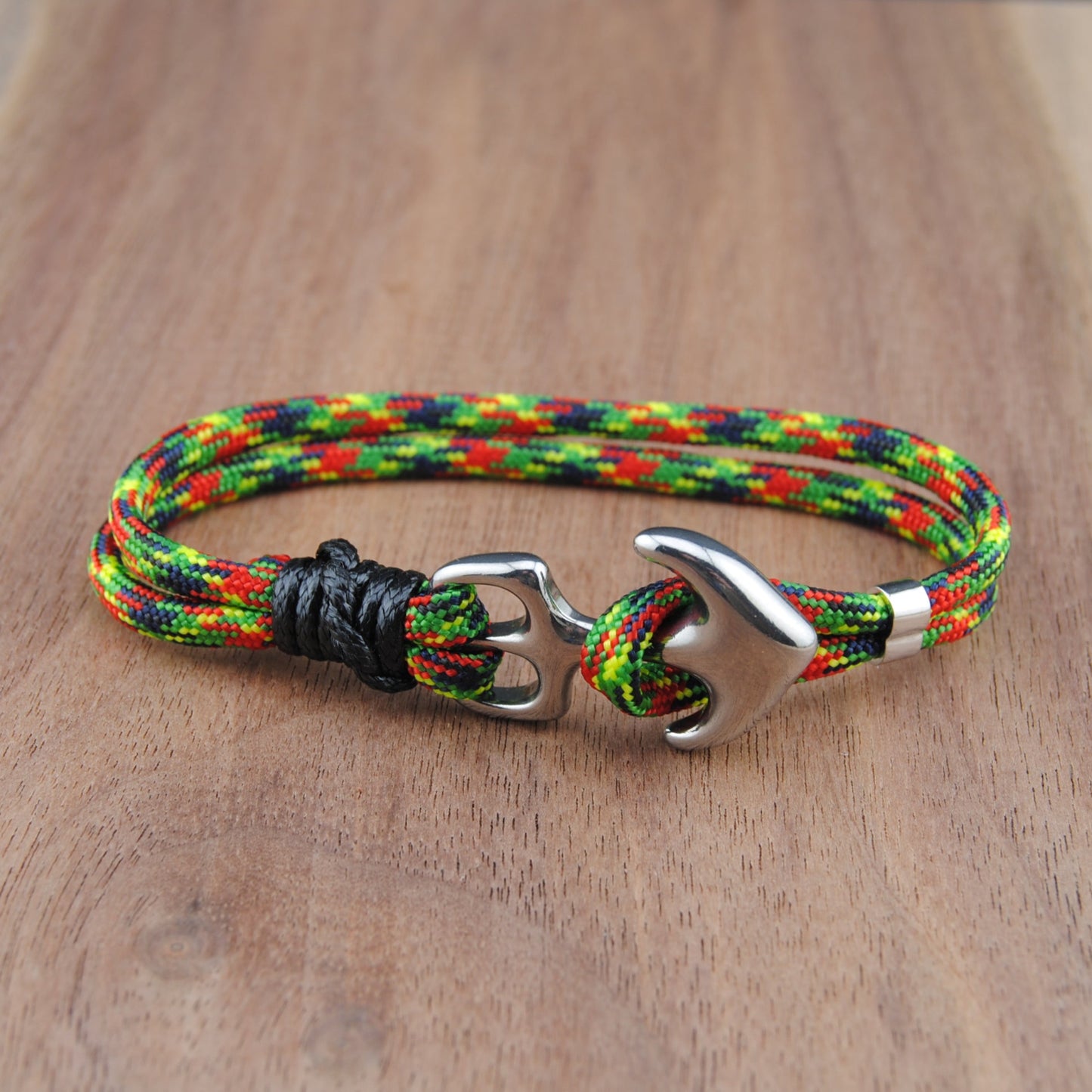 A braided paracord bracelet with red, green, and yellow threads, featuring a black knotted accent and a silver-toned anchor-shaped metal clasp, displayed on a wooden surface.