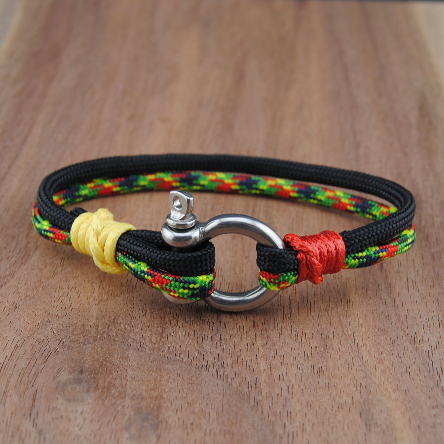 A handmade braided cord bracelet with a silver metal ring clasp, resting on a wooden surface. The bracelet is primarily black with braided accents of yellow, red, and green.
