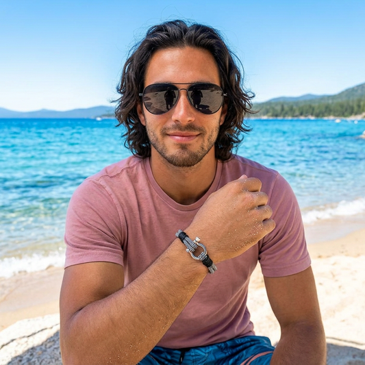 Man wearing sunglasses and a pink shirt sitting on a beach with clear blue water and mountains in the background.