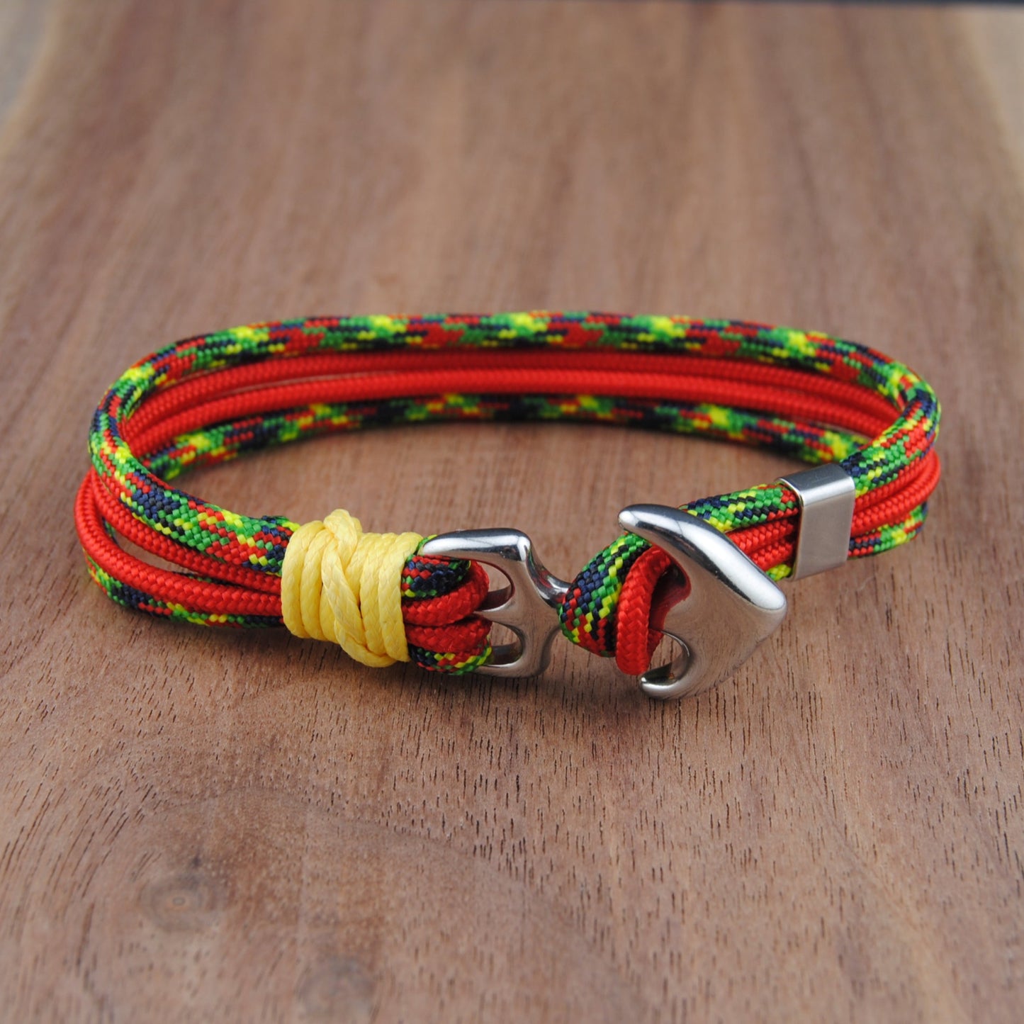 A multi-strand paracord bracelet in red, green, and yellow with a silver metal anchor clasp, resting on a wooden surface.