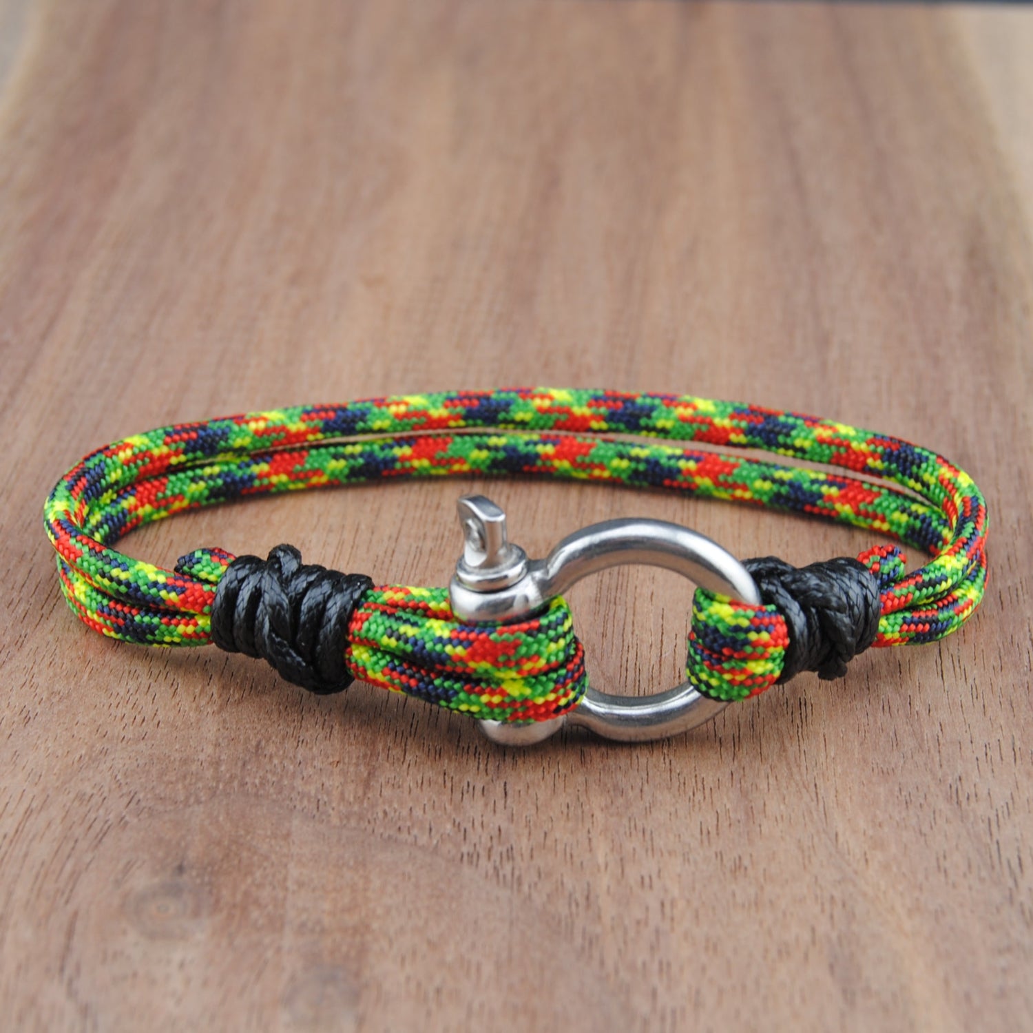 A braided multicolor cord bracelet with red, yellow, green, and black woven strands, featuring a silver metal ring and toggle clasp, resting on a wooden surface.