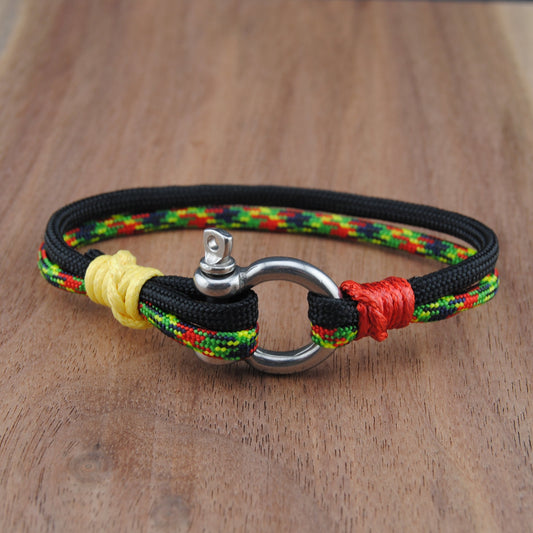 A handmade braided cord bracelet with a silver metal ring clasp, resting on a wooden surface. The bracelet is primarily black with braided accents of yellow, red, and green.