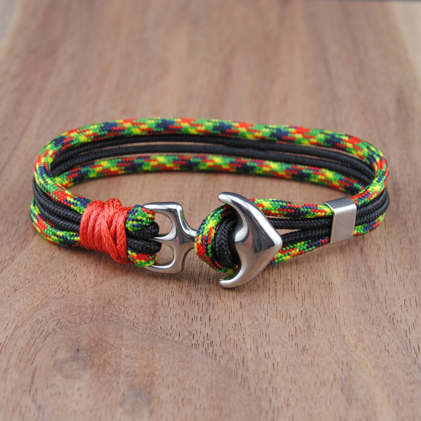 A braided cord bracelet with a silver anchor clasp, resting on a wooden surface. The bracelet features a multicolor pattern of black, green, yellow, and red cords, accented by a bright red wrap near the clasp.