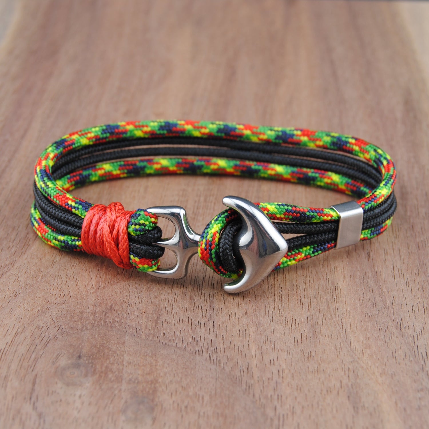 A braided cord bracelet with a silver anchor clasp, resting on a wooden surface. The bracelet features a multicolor pattern of black, green, yellow, and red cords, accented by a bright red wrap near the clasp.